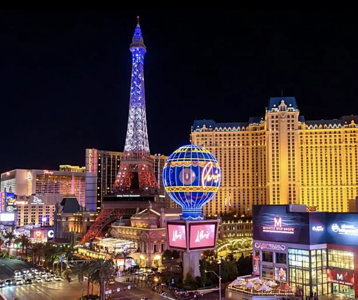 Las Vegas strip at night, showcasing the illuminated replica of the Eiffel Tower at Paris Las Vegas hotel, with surrounding buildings aglow with neon signs. It's a spectacle reminiscent of a grand Spring Gathering under the vibrant desert sky. - Intercoiffure Canada America