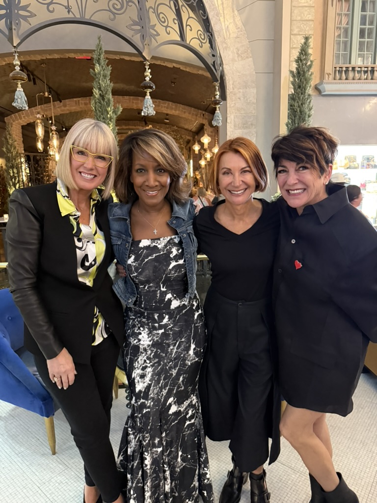 Four women stand closely together indoors, smiling at the camera. The setting features elegant decor with arches, greenery, and hanging lights in the background. - Intercoiffure Canada America
