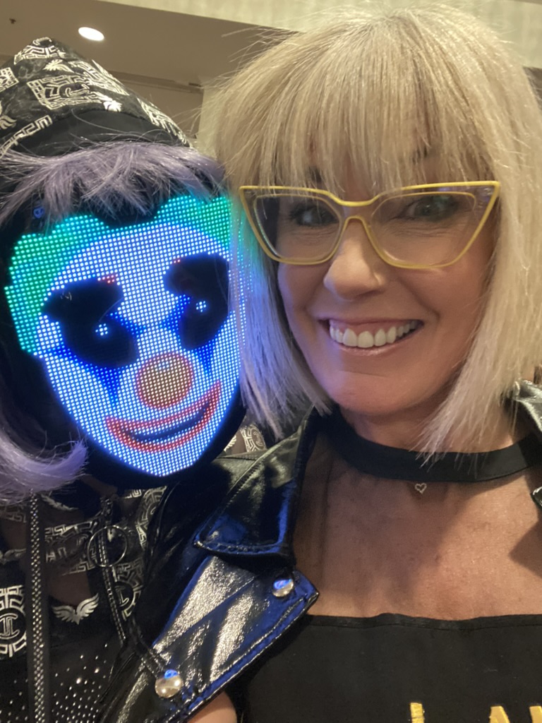 Two people pose for a photo; one wears a hooded jacket and a glowing LED mask with a colorful face, the other has blonde hair, yellow glasses, and a black jacket. - Intercoiffure Canada America