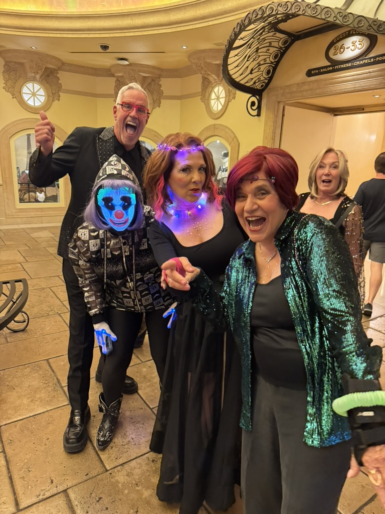 A group of five adults in costumes pose indoors; one wears glowing face paint, another has LED necklace, others are dressed in party attire with sequins and accessories. - Intercoiffure Canada America