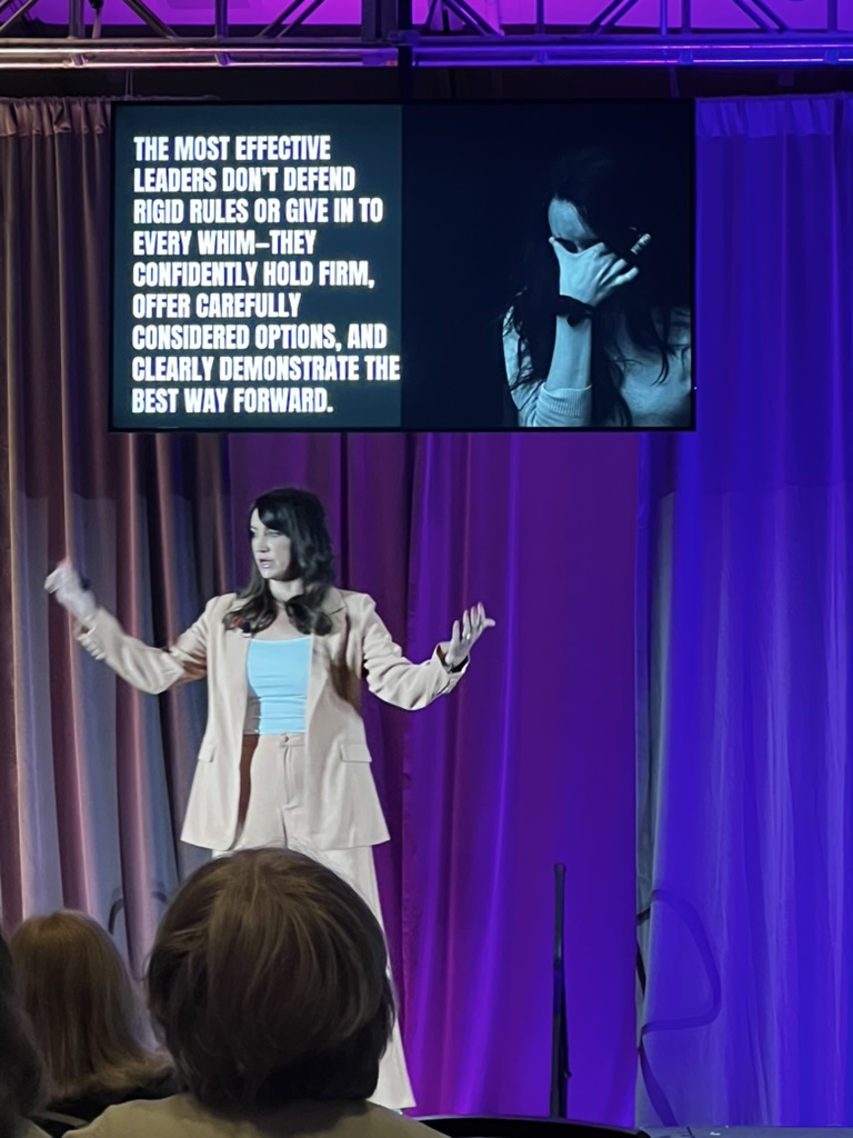 A woman in a light suit speaks onstage under a screen displaying a quote about effective leadership and a black-and-white photo of a person with a hand covering their face. - Intercoiffure Canada America