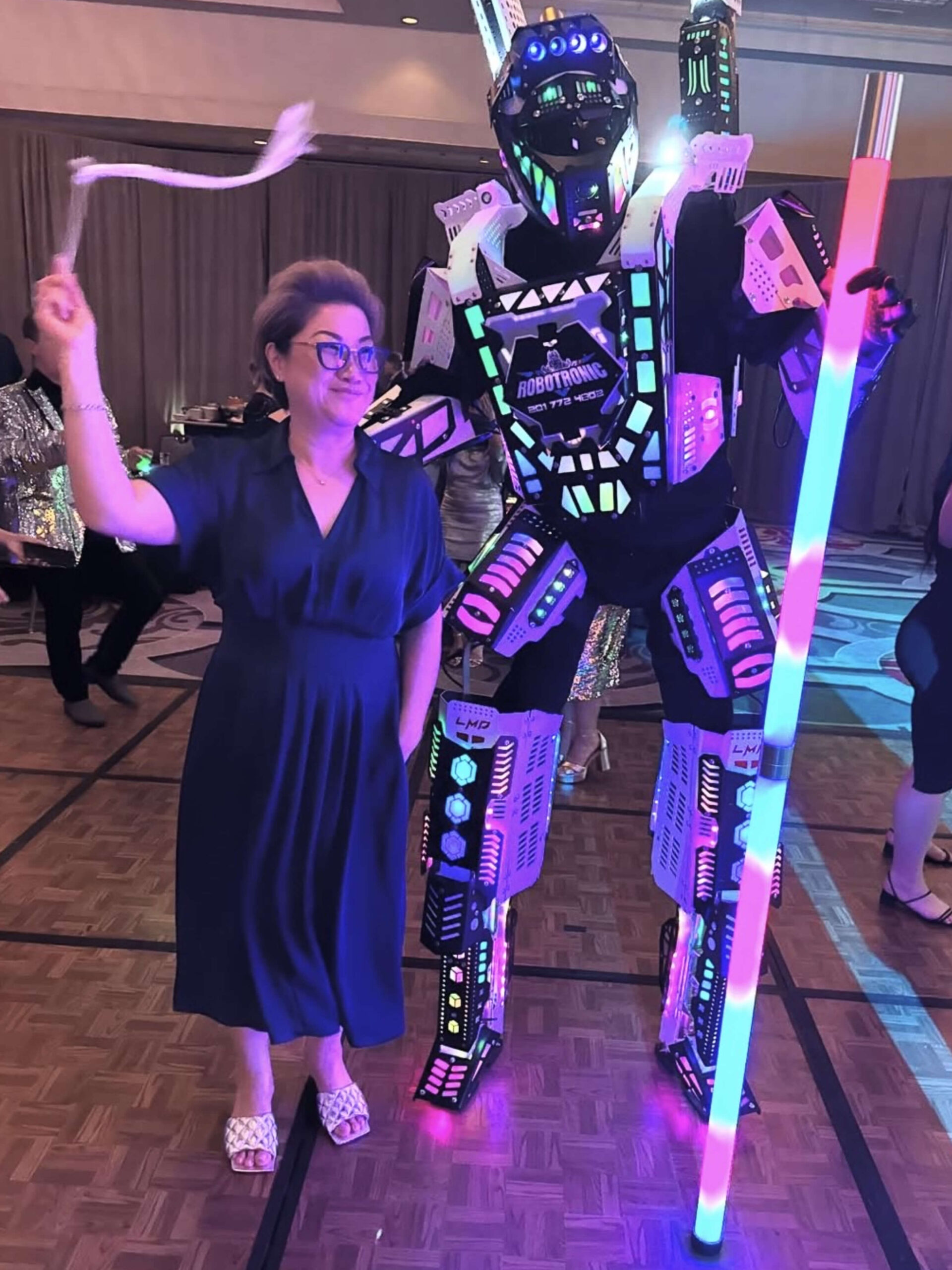 At the ICA Spring Gathering Guides event, a woman in a blue dress stands beside a person in an illuminated robot costume holding a glowing staff on the dance floor. - Intercoiffure Canada America