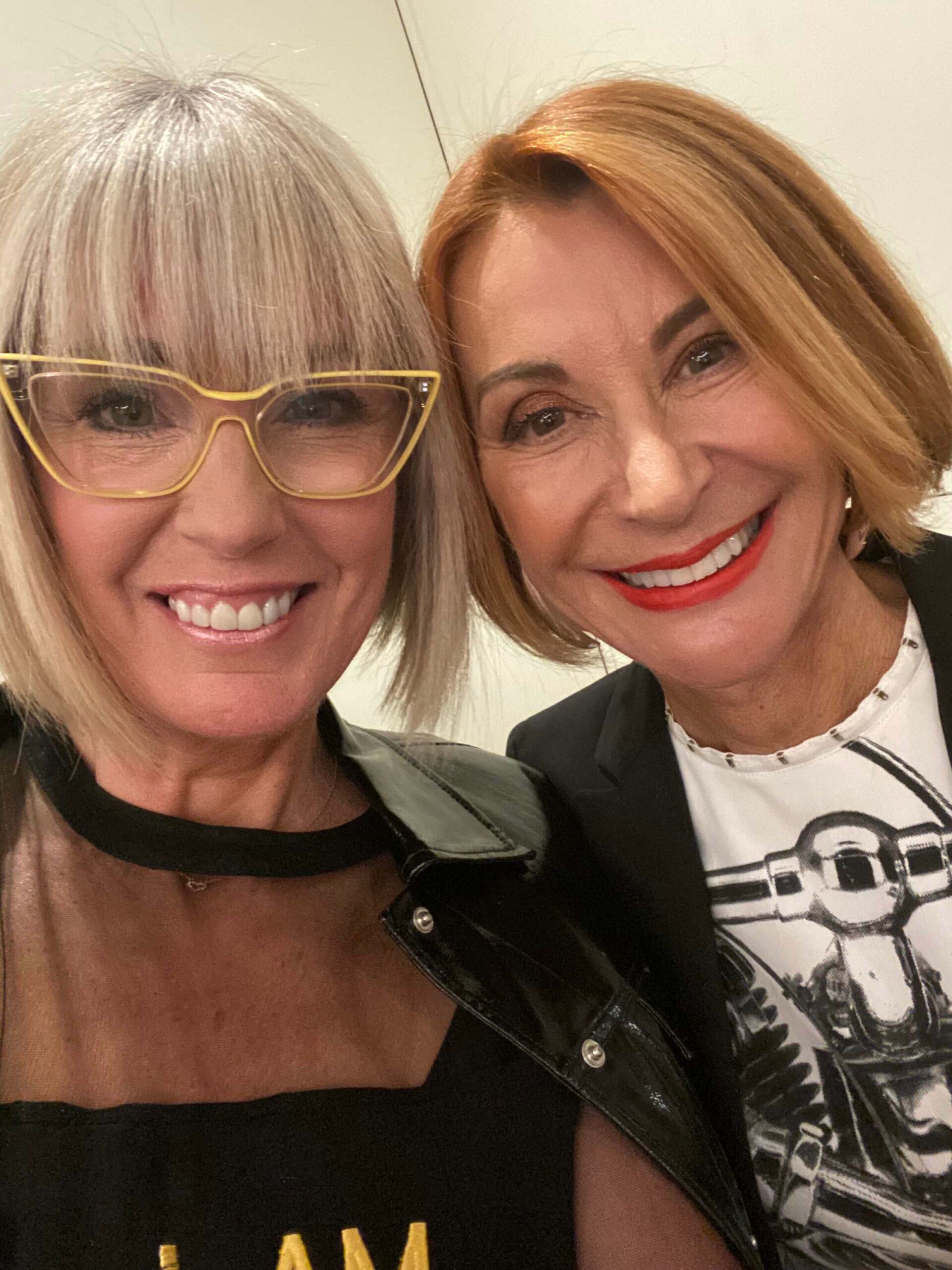 Two women with short hair, glasses, and bright lipstick smiling together for a close-up selfie. - Intercoiffure Canada America