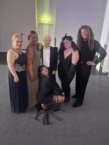 Six people pose together indoors, dressed in formal and stylish evening attire, celebrating 100 Years of Intercoiffure Mondial. White walls and a closed door with a lit exit sign are visible in the background. - Intercoiffure Canada America