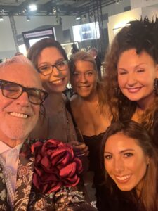 Five people, including ICA Members, smiling and posing for a selfie at an indoor event; one man in a floral jacket with a large red flower, and four women, one with a striking black headpiece, celebrating Intercoiffure Mondial’s 100 Years. - Intercoiffure Canada America