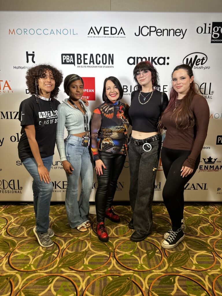 Five women stand together smiling in front of a sponsor step-and-repeat banner at an Intercoiffure Salons event, showcasing the perfect match of talent and style. - Intercoiffure Canada America