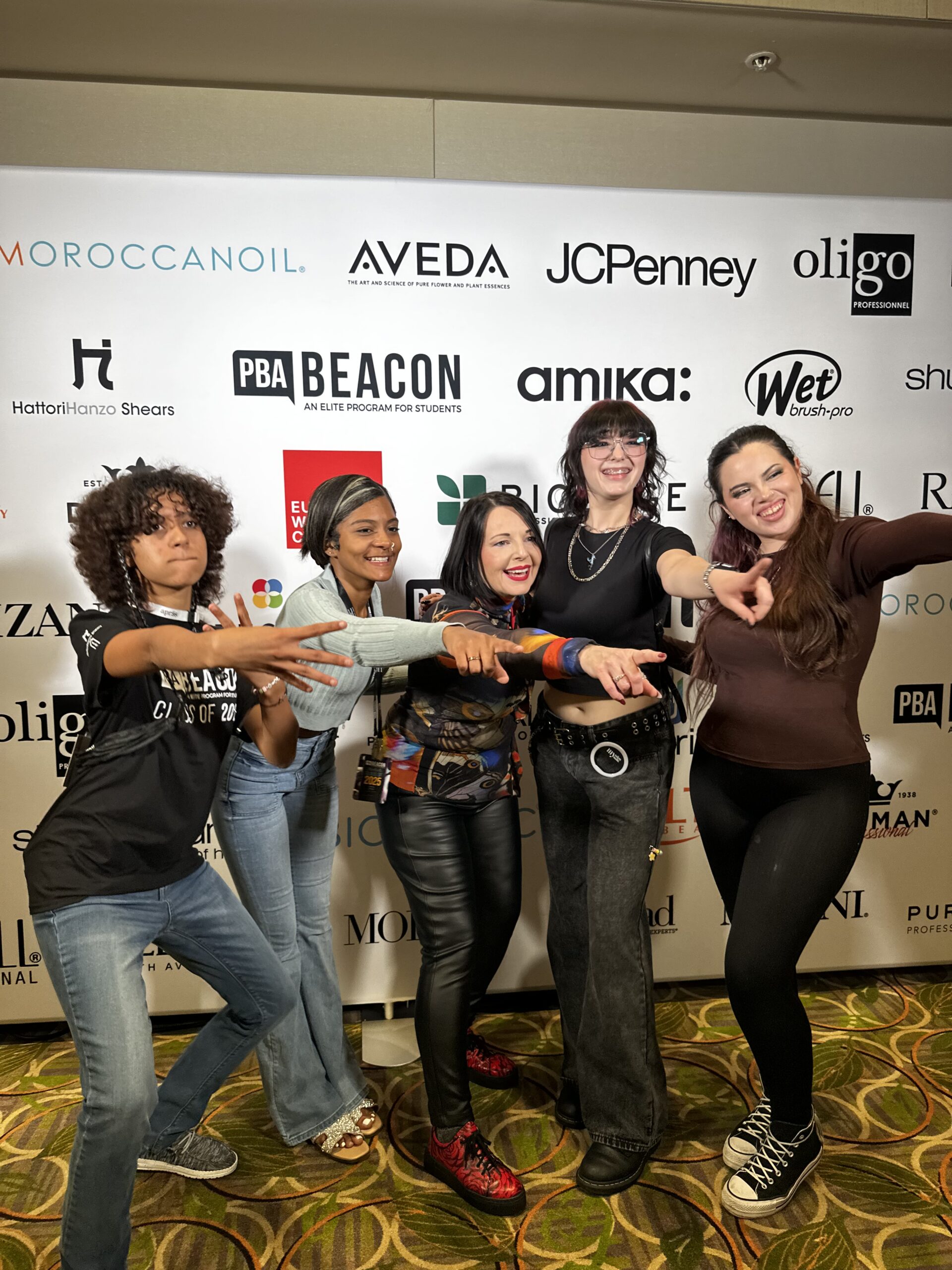 Five women stand together in front of a sponsor logo wall, smiling and making playful hand gestures toward the camera, celebrating their Perfect Match at Intercoiffure Salons. - Intercoiffure Canada America