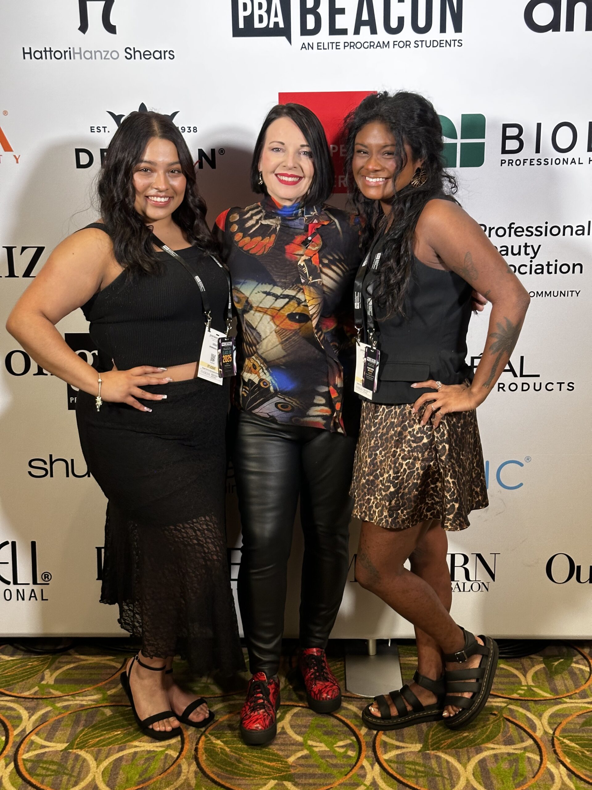 Three women stand together smiling in front of a step-and-repeat banner at a professional event for Intercoiffure Salons, wearing black and patterned outfits with event lanyards—a perfect match of style and professionalism. - Intercoiffure Canada America