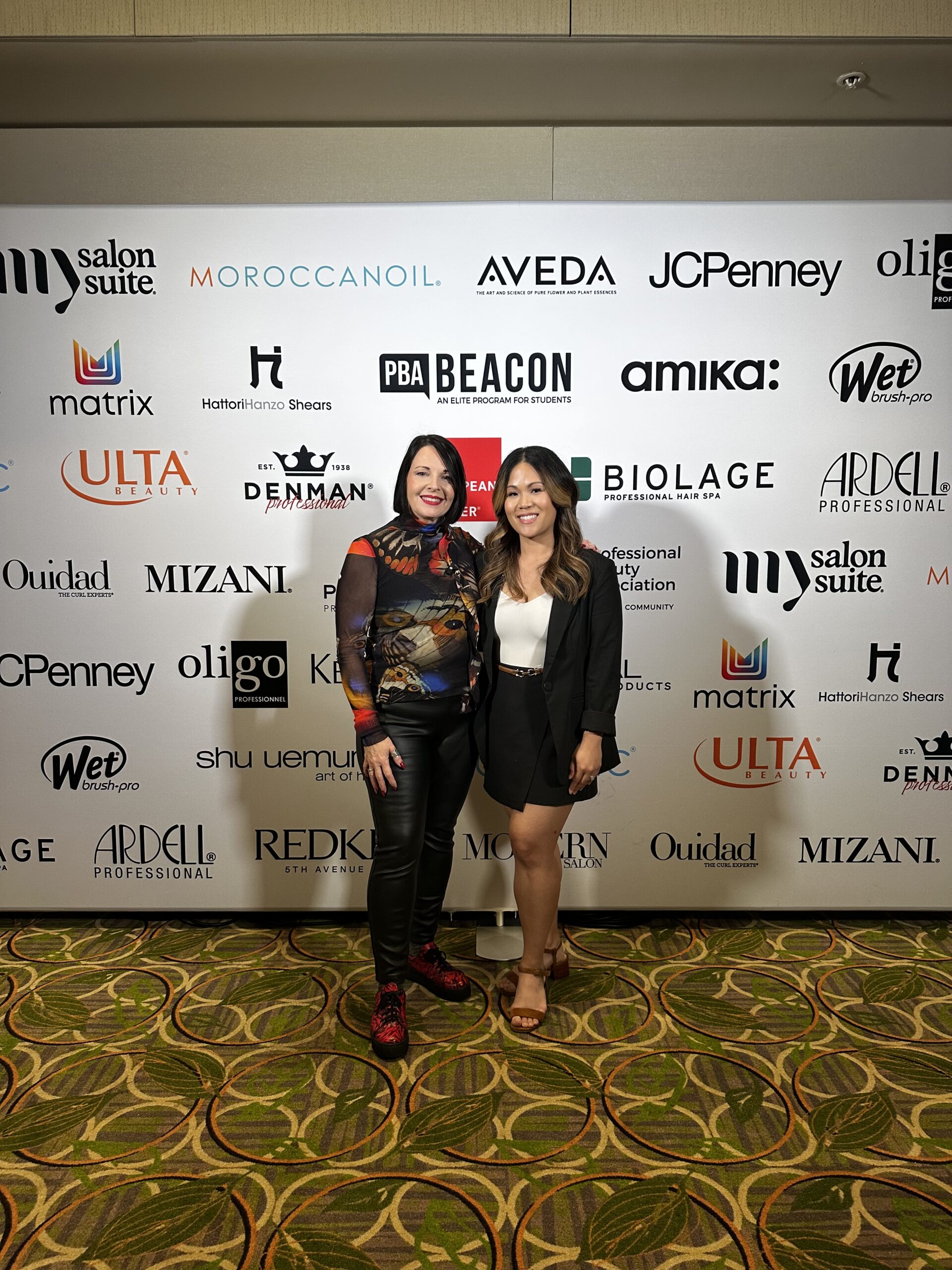 Two women stand side by side, smiling, in front of a step-and-repeat banner featuring various beauty and salon brand logos, including Intercoiffure Salons—perfect for inspiring future Beacon Students. - Intercoiffure Canada America