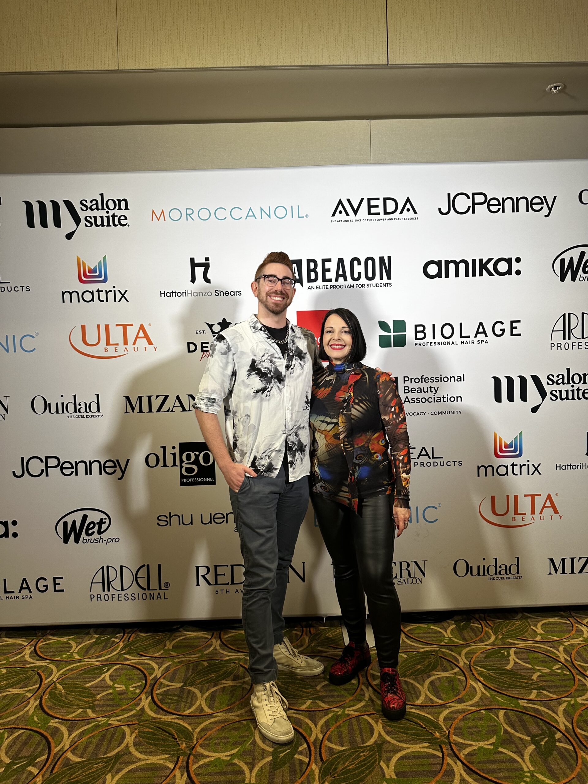 Two people stand smiling in front of a step-and-repeat banner featuring Intercoiffure Salons and other beauty brand logos on a patterned carpet, showing they're a perfect match for the industry. - Intercoiffure Canada America