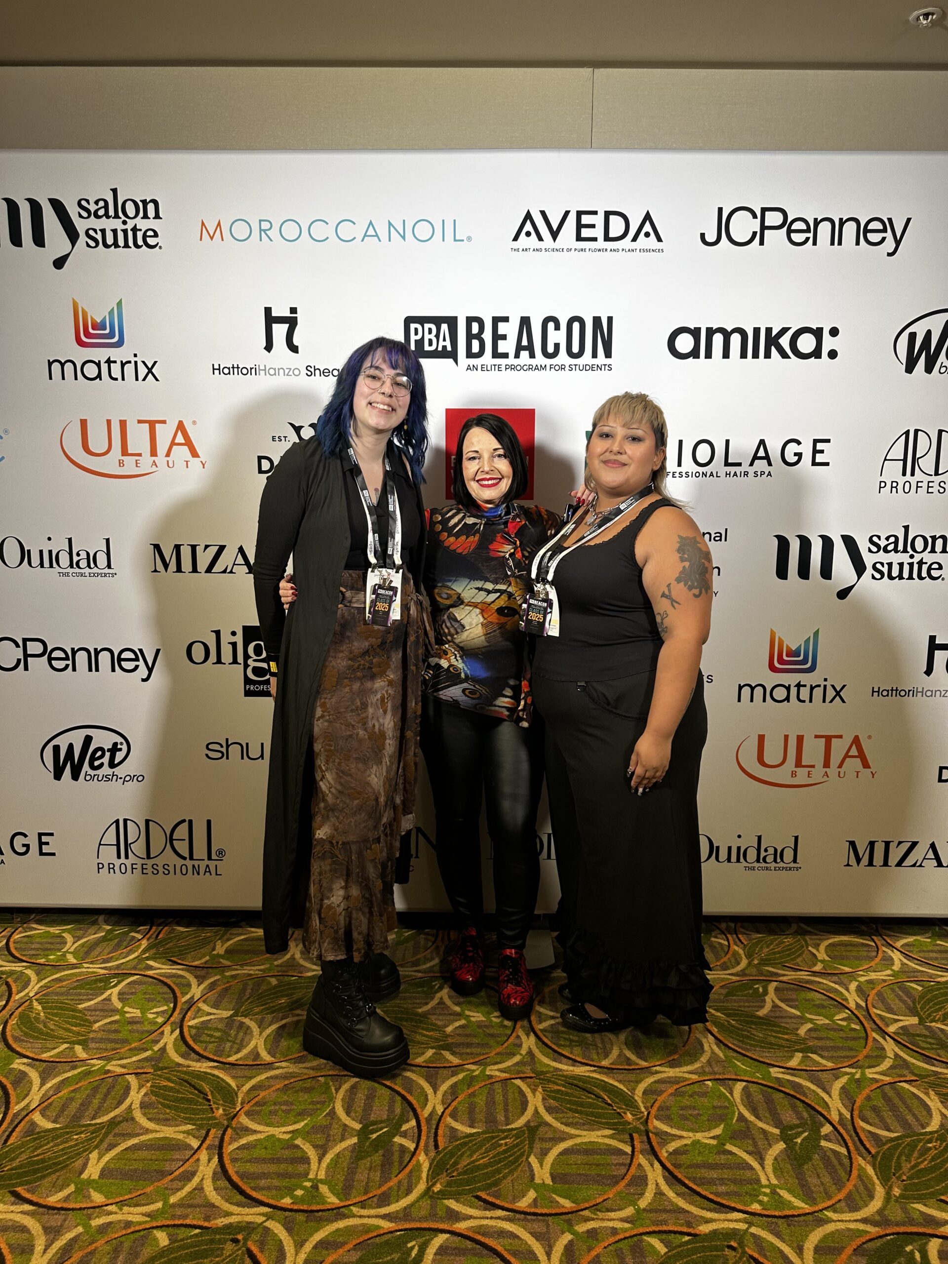 Three women pose together in front of a sponsor backdrop at an event, wearing Beacon Students conference badges and casual attire. - Intercoiffure Canada America