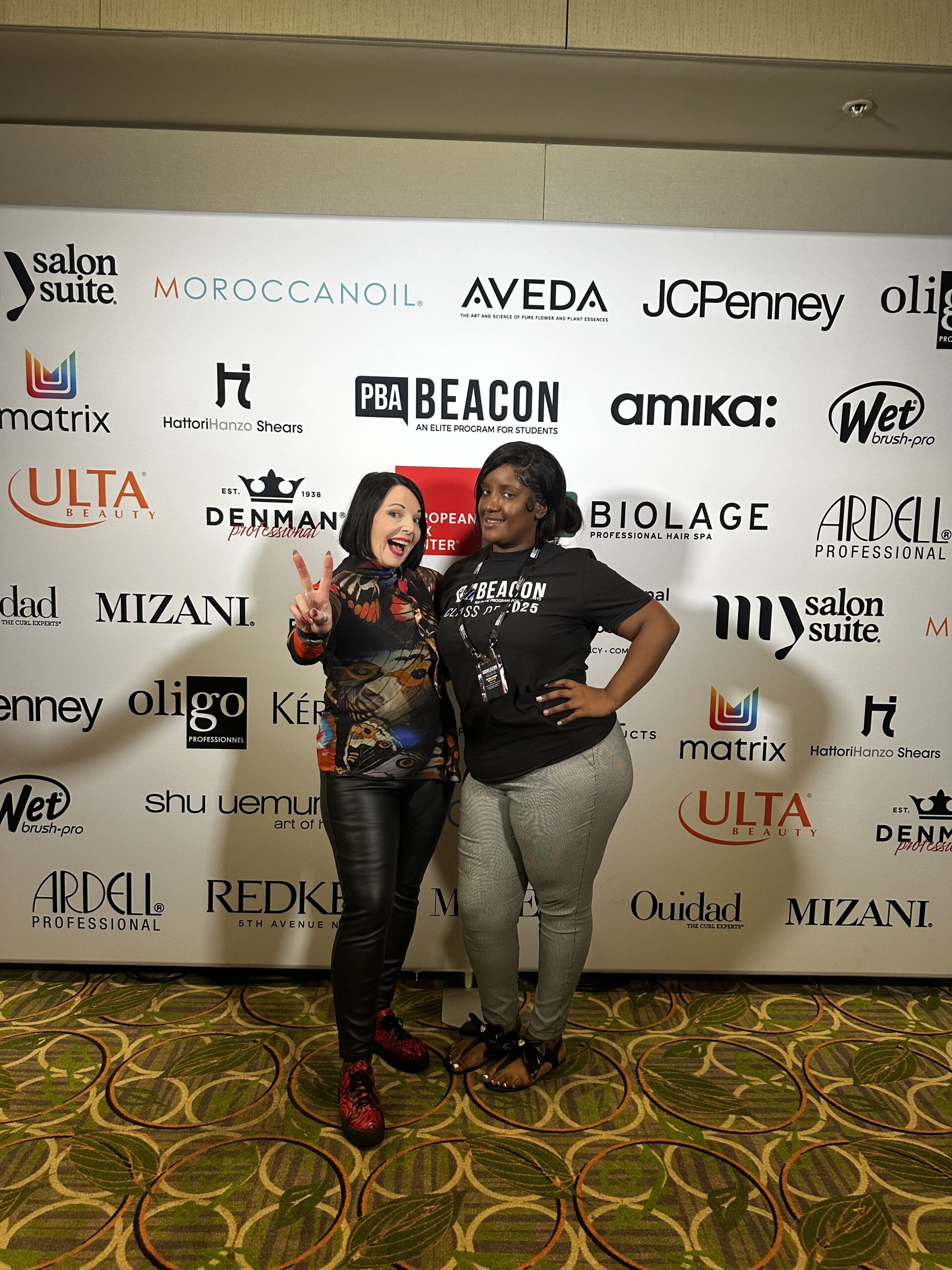 Two women, Beacon Students, pose and smile in front of a step-and-repeat banner featuring various beauty and haircare brand logos, including Intercoiffure Salons, at an indoor event. - Intercoiffure Canada America