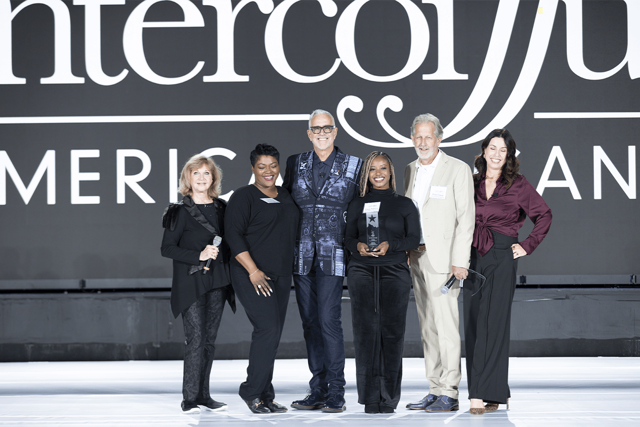 Six people stand on stage smiling, holding a trophy, in front of a large graphic background with bold text. They are dressed in formal and semi-formal attire. - Intercoiffure Canada America - Intercoiffure Canada America - Intercoiffure Canada America - Intercoiffure Canada America - Intercoiffure Canada America