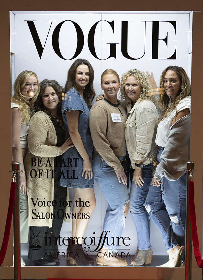 Six women pose and smile inside a photo booth styled as a Vogue magazine cover, with red ropes and salon-related text featured on the display. - Intercoiffure Canada America - Intercoiffure Canada America - Intercoiffure Canada America - Intercoiffure Canada America - Intercoiffure Canada America - Intercoiffure Canada America