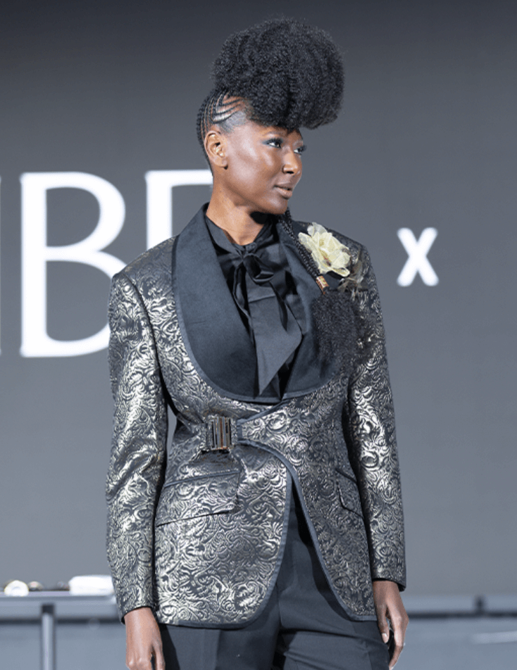 A person wearing a patterned metallic suit jacket, black shirt with a bow, and floral boutonniere stands in front of a blurred background with large white letters. - Intercoiffure Canada America - Intercoiffure Canada America - Intercoiffure Canada America - Intercoiffure Canada America