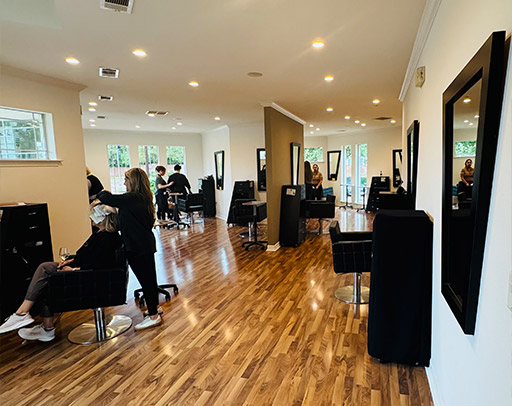 A modern hair salon with wooden floors, black chairs, large mirrors, and several people getting haircuts and styling—there’s a touch of McGaha Magic in the air, perfect for a 2026 Spring Regional Preview. - Intercoiffure Canada America