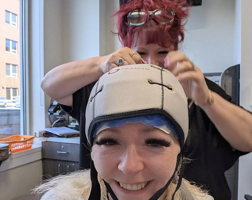 A woman smiles while wearing a cooling cap, as another person offers support by adjusting the straps in a clinical setting—providing comfort for cancer patients. - Intercoiffure Canada America