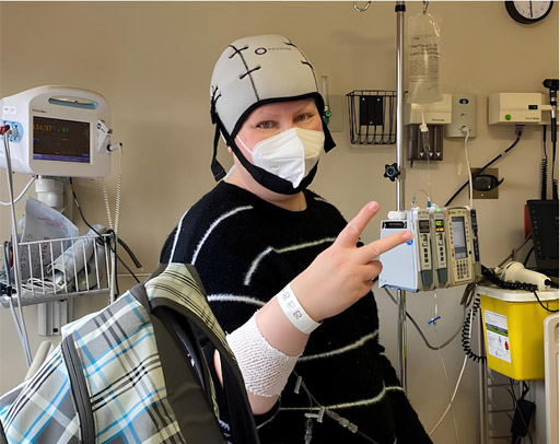 Person wearing a striped sweater, cooling cap, and mask sits in a medical setting, giving a peace sign—a moment of strength shared by many cancer patients. Medical equipment and monitors are visible in the background. - Intercoiffure Canada America