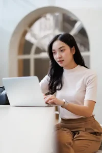 A woman sits at a table using a laptop computer, with a bag beside her and large window in the background. - Intercoiffure Canada America