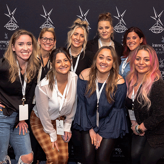 Eight women wearing conference badges pose and smile in front of a branded backdrop at the Spring Gathering 2026, capturing the spirit of this professional event. - Intercoiffure Canada America
