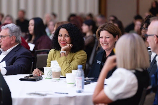 A group of people sit at round tables, attentively listening and smiling during an indoor conference or seminar. - Intercoiffure Canada America