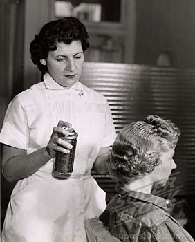 A woman in a white uniform uses beauty technology as she sprays hairspray on another woman's styled hair in a salon. - Intercoiffure Canada America