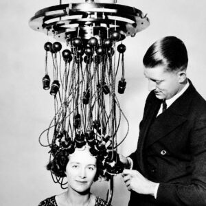 A woman sits beneath an early 20th-century perm machine, its wires a testament to the technology bringing major change to beauty routines, while a man attentively adjusts the device. - Intercoiffure Canada America