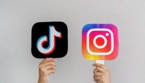 Two hands hold up signs displaying the TikTok logo on the left and the Instagram logo on the right, highlighting a major change in beauty and technology trends against a plain light background. - Intercoiffure Canada America