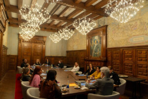 People sit around a large conference table in an ornate room with wood paneling, chandeliers, and wall art, engaged in a formal meeting or discussion of salon leadership and the future of the salon industry. This distinguished gathering could represent ICA’s NEXT Salon Leaders exchanging ideas and shaping new directions for their field. - Intercoiffure Canada America