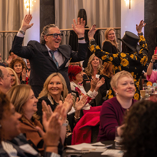 A man in a suit and a person in a floral jacket stand with raised arms as an audience applauds in a crowded, warmly lit event room. - Intercoiffure Canada America