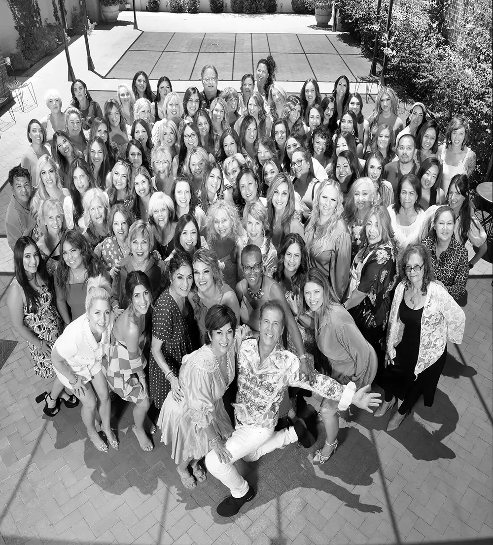 Large team of people posing together outdoors on a paved courtyard, all facing the camera and smiling. The image is in black and white. - Intercoiffure Canada America